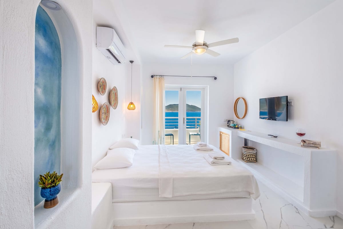A hotel room with a bed, a ceiling fan, a TV, a window with a view of the sea, and a small plant.