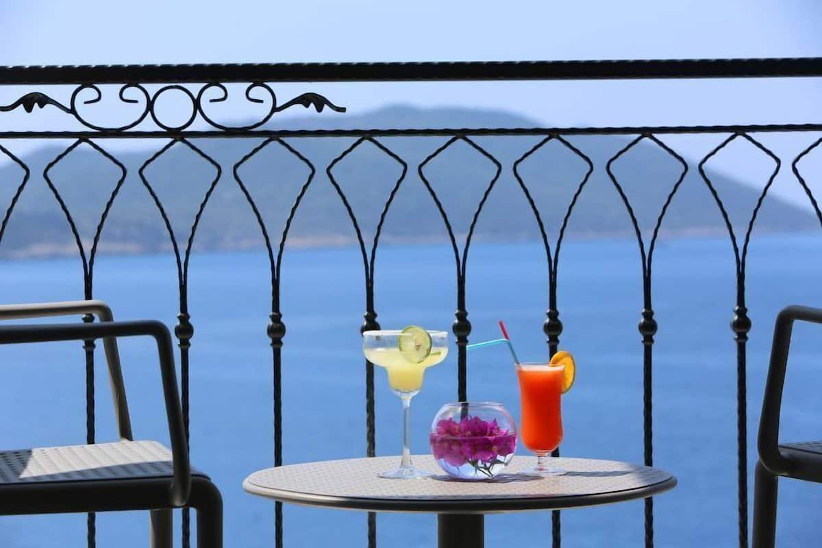 A round table with two drinks and a flower vase on a balcony with a metal railing and a view of the sea.