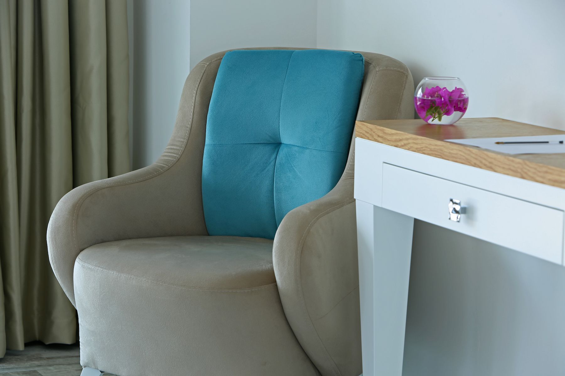 A beige armchair with a teal cushion, a white desk with a wooden top, and a glass vase with purple flowers.