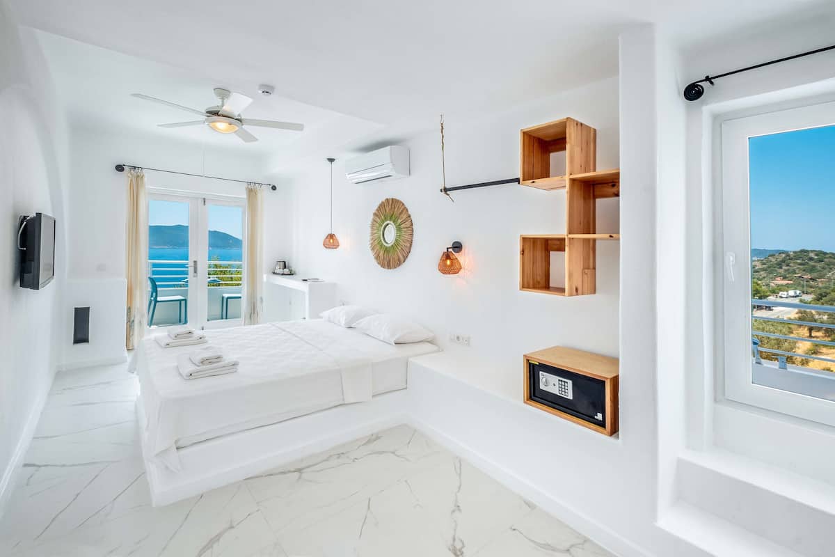 A modern bedroom with a bed, a ceiling fan, a TV, a wall-mounted shelf, and a view of the sea.