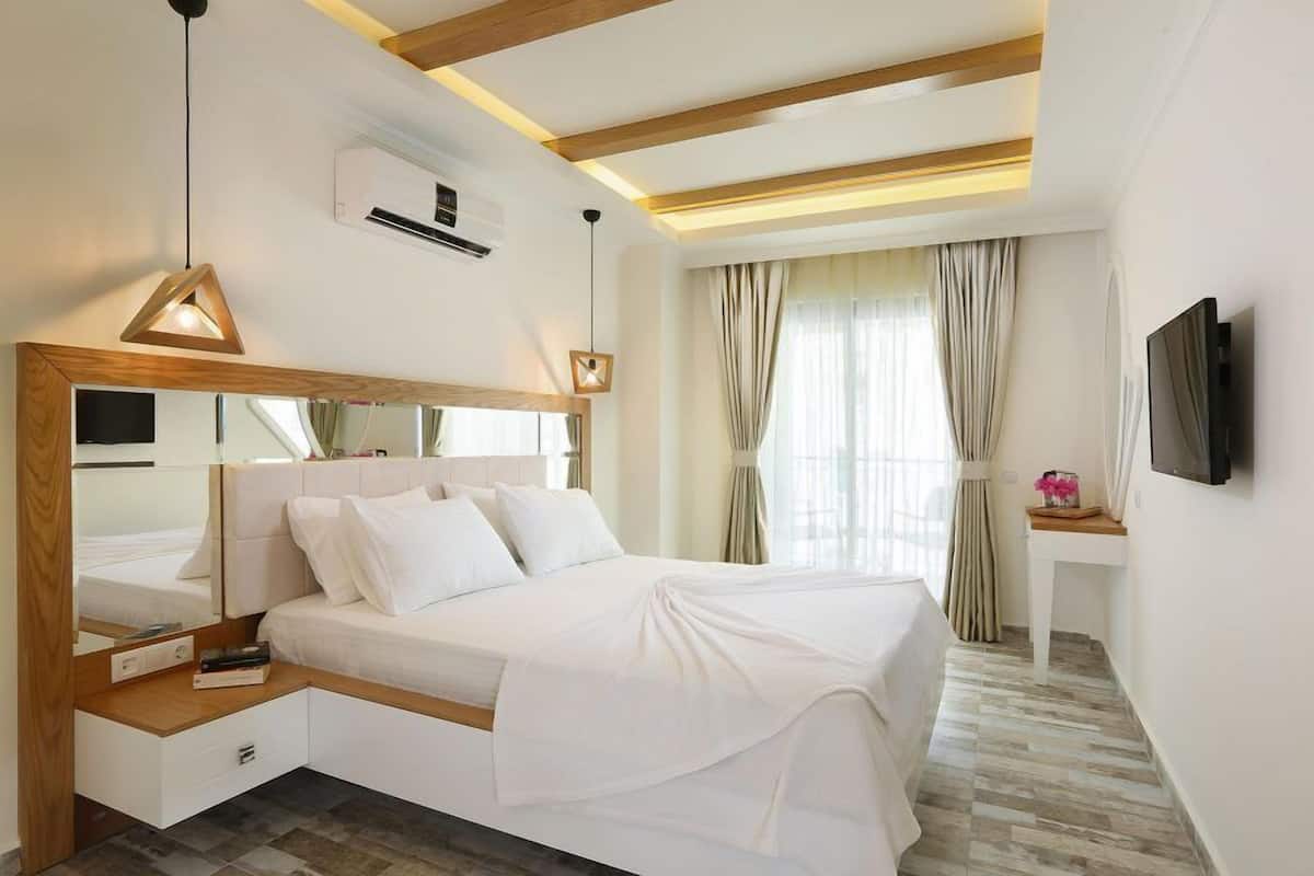 A modern hotel room with a large bed, a bedside table, a mirror, a TV mounted on the wall, and a window with curtains.
