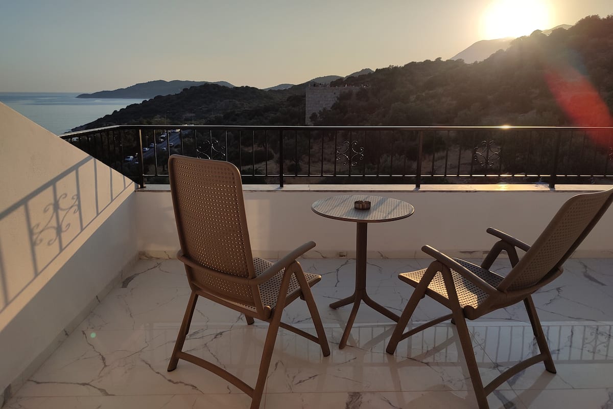 A balcony with two chairs and a small table, overlooking a coastal landscape with mountains and the sea.