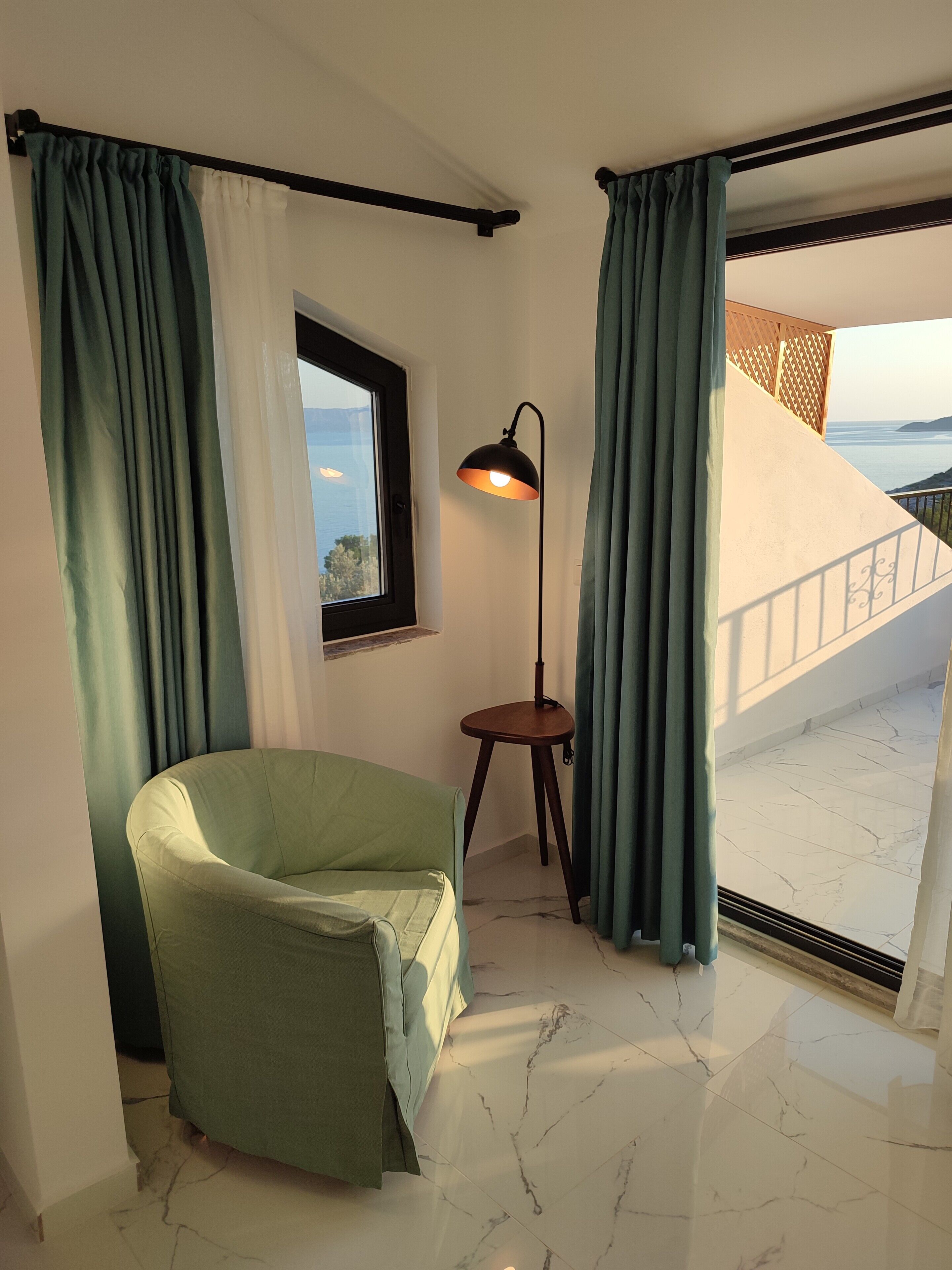 Una stanza con una poltrona verde, una lampada da terra, una finestra vista mare e un balcone con ringhiera.