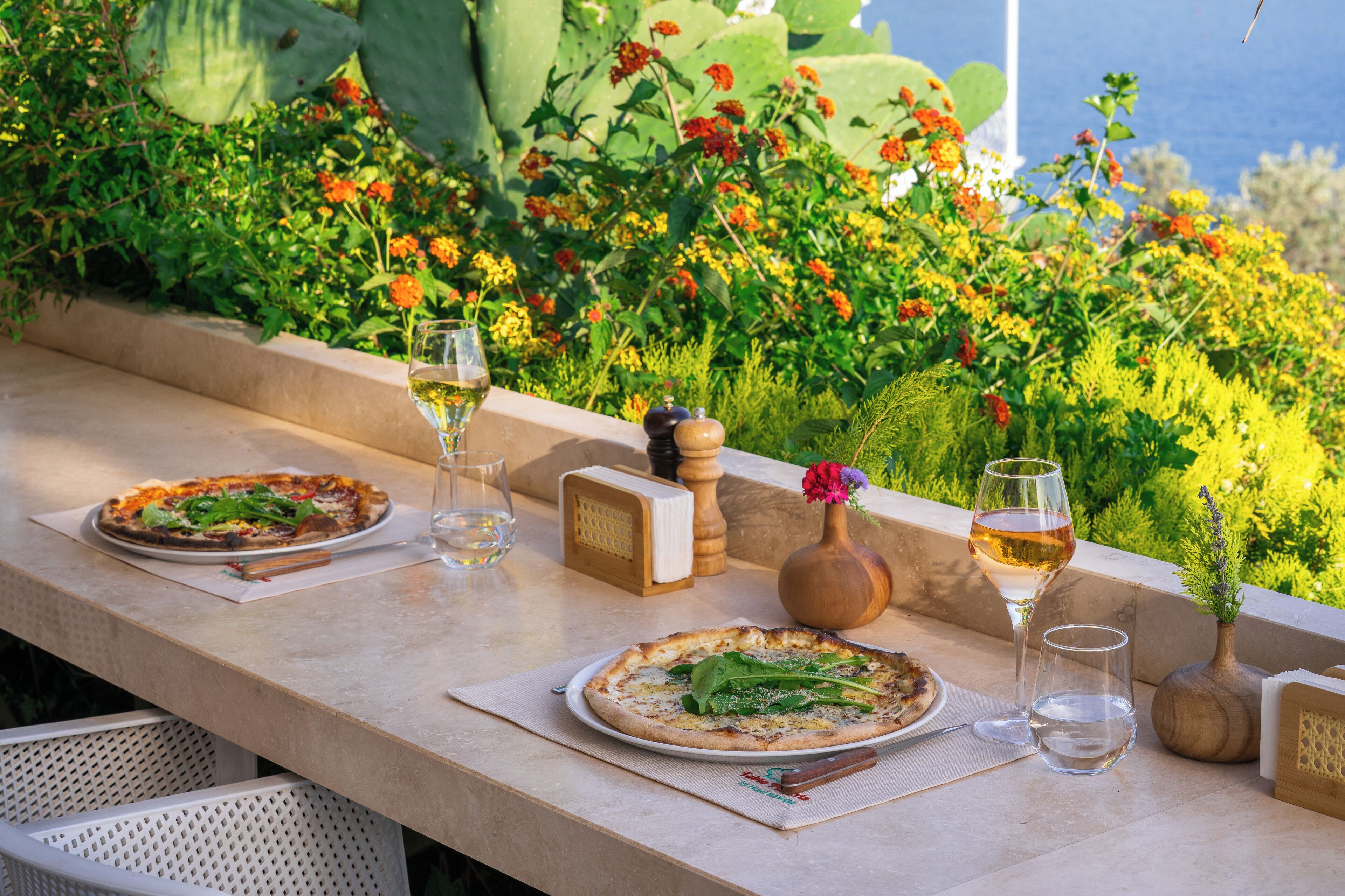 Una mesa con dos pizzas, una copa de vino y un florero con flores.