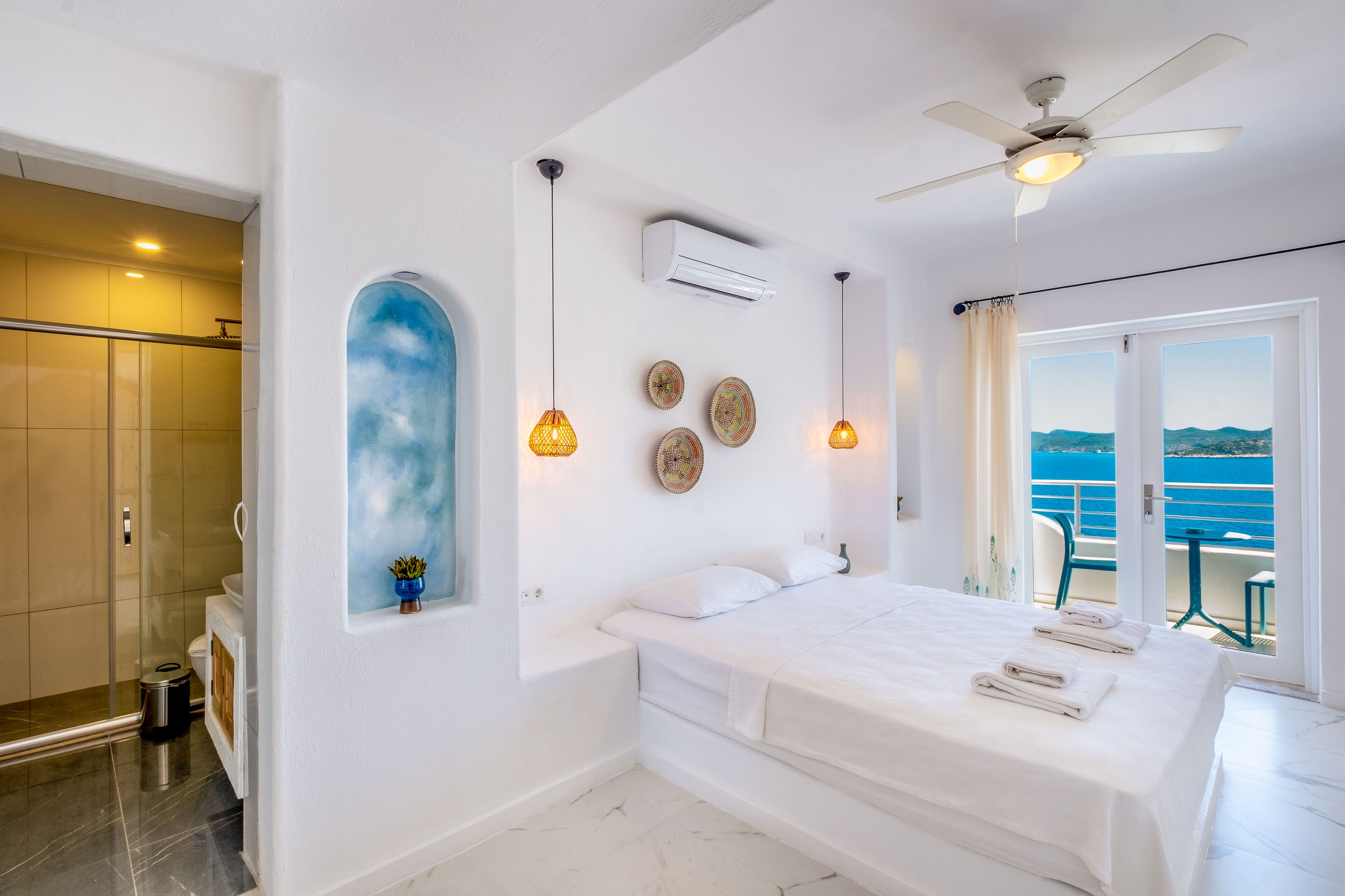 A modern bedroom with a large bed, a ceiling fan, and a view of the sea.