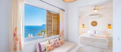 A room with a large window overlooking the sea, a sofa with patterned cushions, a hanging light fixture, and a ceiling fan.