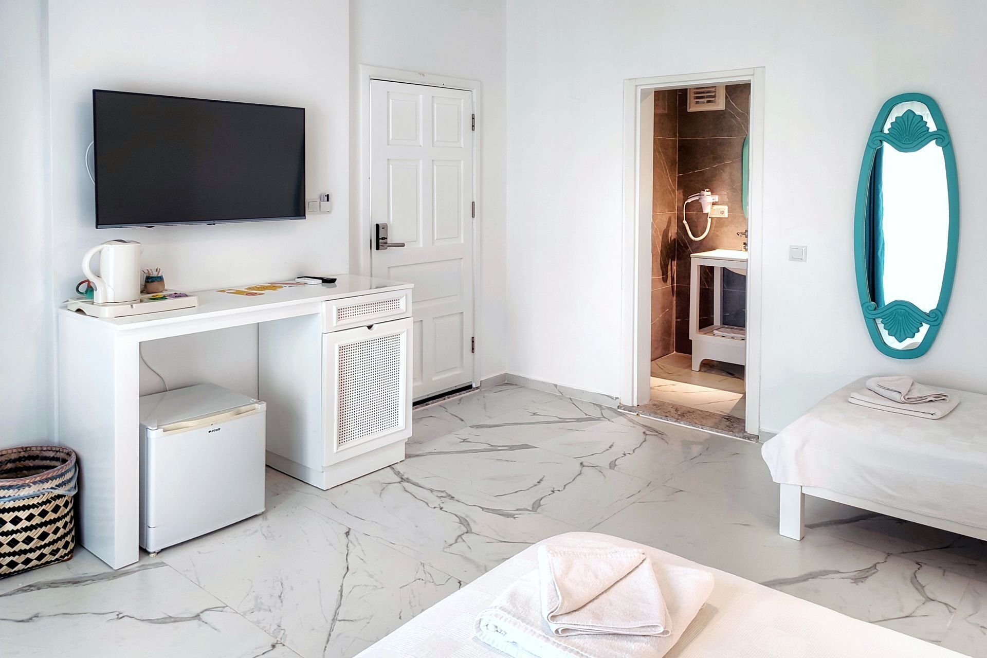 A hotel room with a flat-screen TV, a desk with a coffee maker, a small fridge, a washing machine, a bathroom with a shower and a mirror, and a bed.