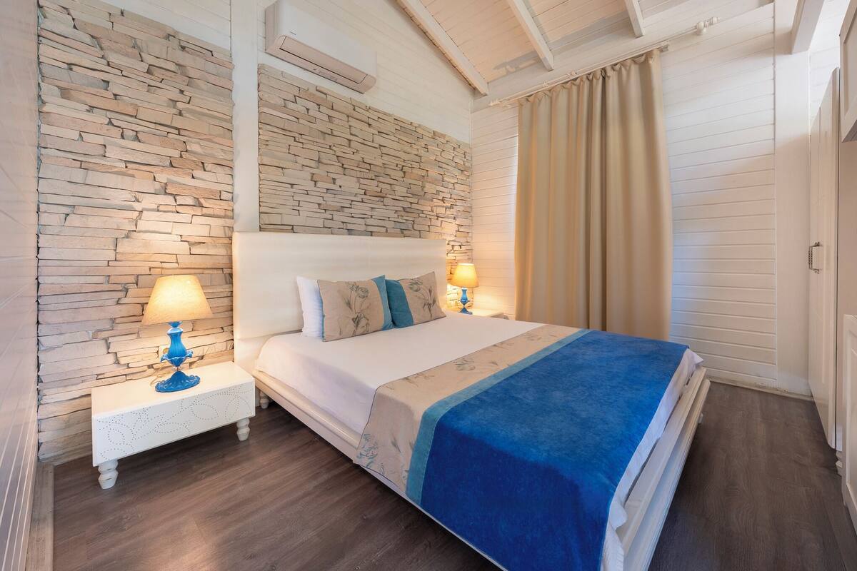 Un dormitorio con pared de piedra, una cama con manta azul, dos lámparas de noche y suelo de madera.