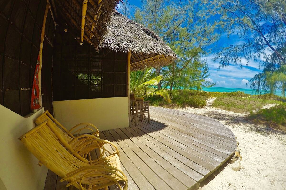 Una terraza de madera con techo de paja, una silla de mimbre y vistas a la playa y el océano.