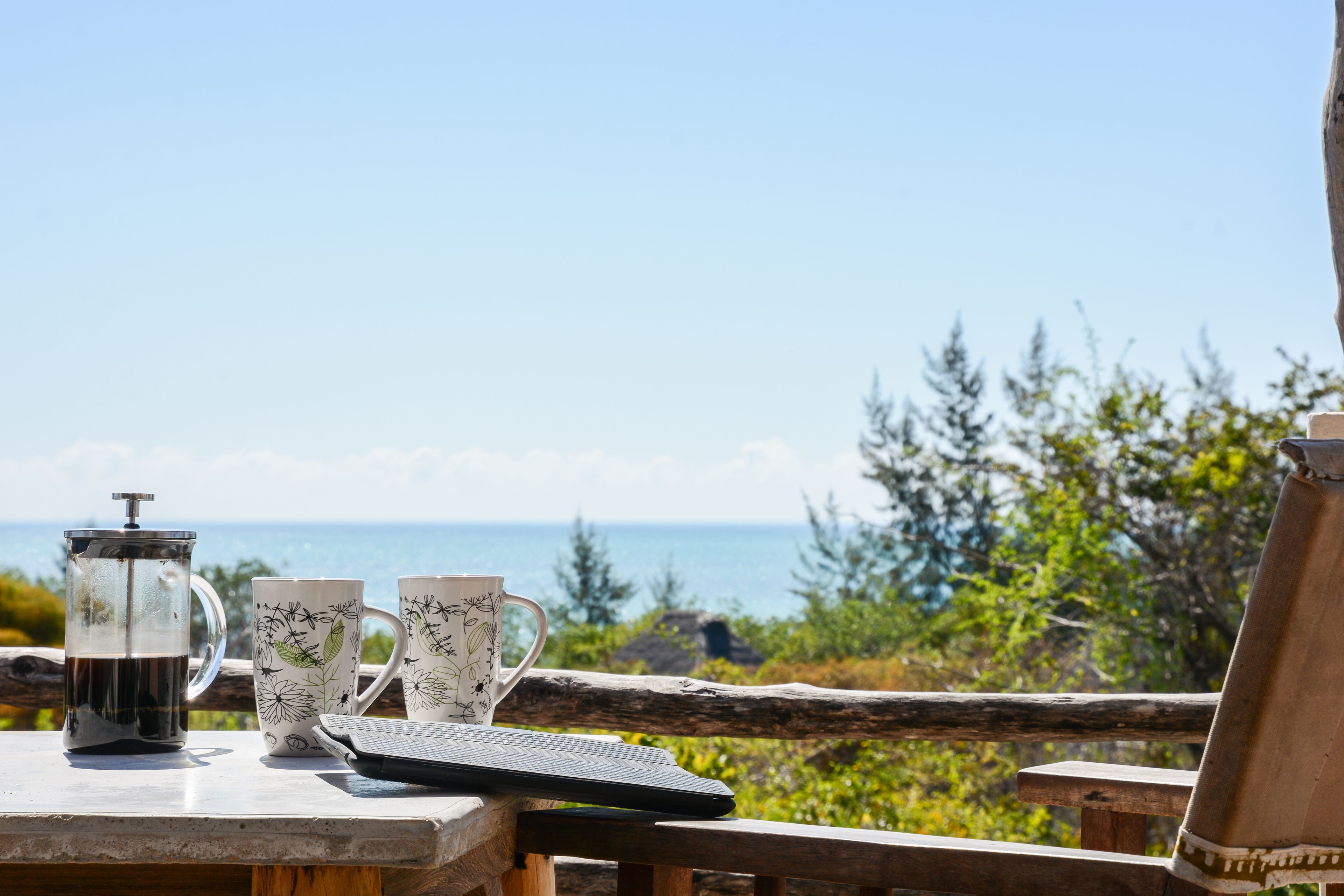 A table with a coffee maker, two patterned mugs, and a smartphone on a wooden deck overlooking a scenic view.