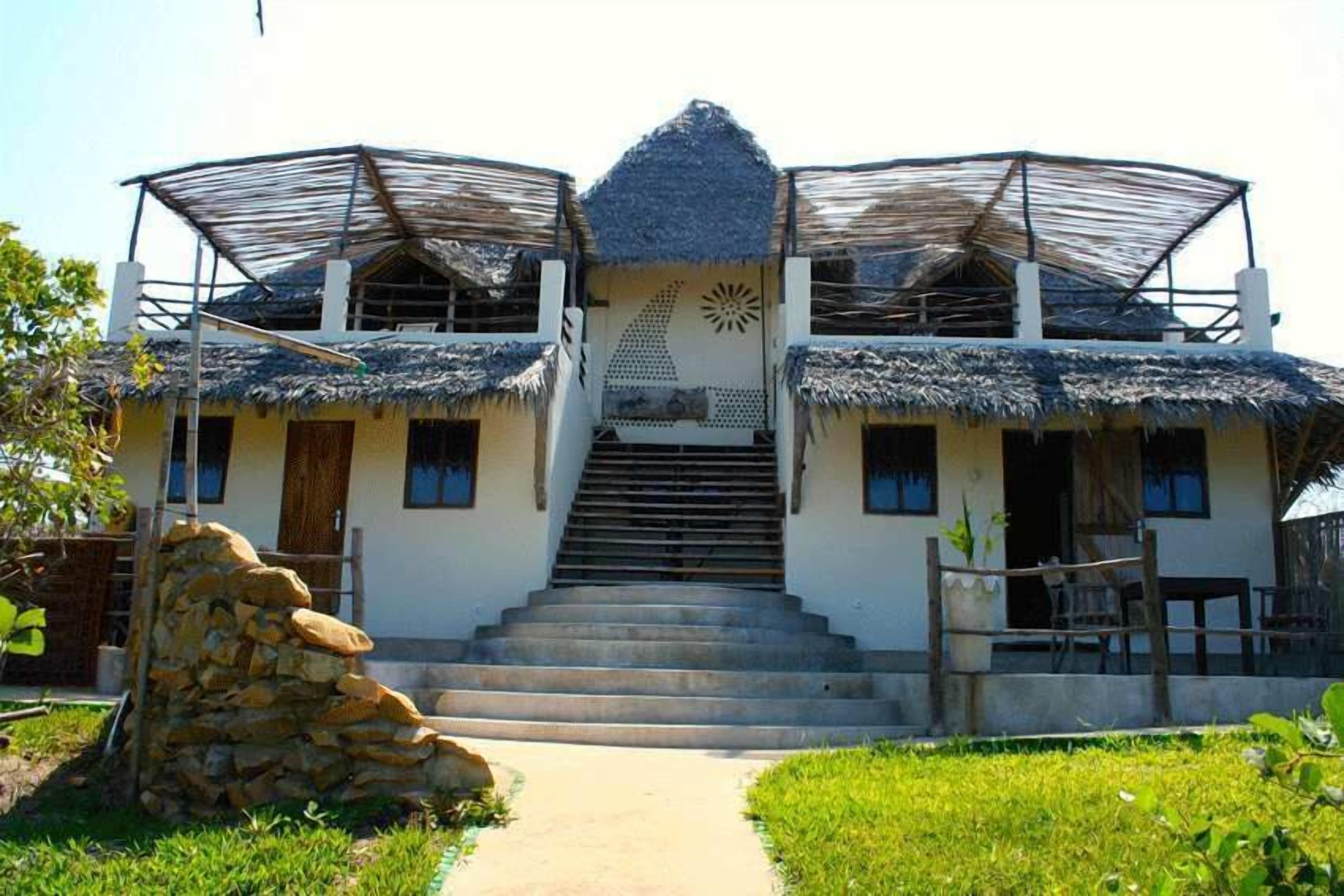 A two-story house with a thatched roof, a staircase leading to the entrance, and a stone wall in the foreground.