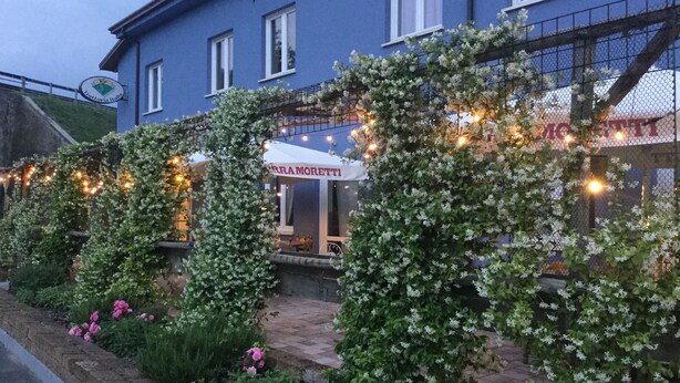 A blue building with a white sign reading "FERRA MORETTI" and a garden with flowering plants and string lights. - Albergo Trattoria Italia (Desenzano del Garda)