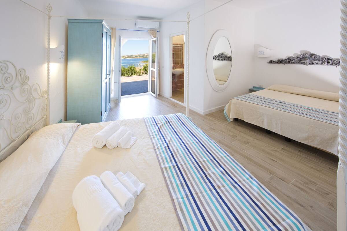 A hotel room with a bed, a bedside table, a mirror, and a view of the sea.
