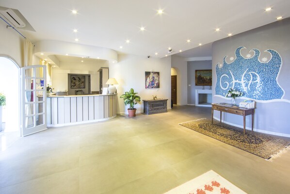 A spacious lobby with a reception desk, a potted plant, a decorative rug, and a wall with a blue abstract design. - Grand Hotel Palau (Palau)