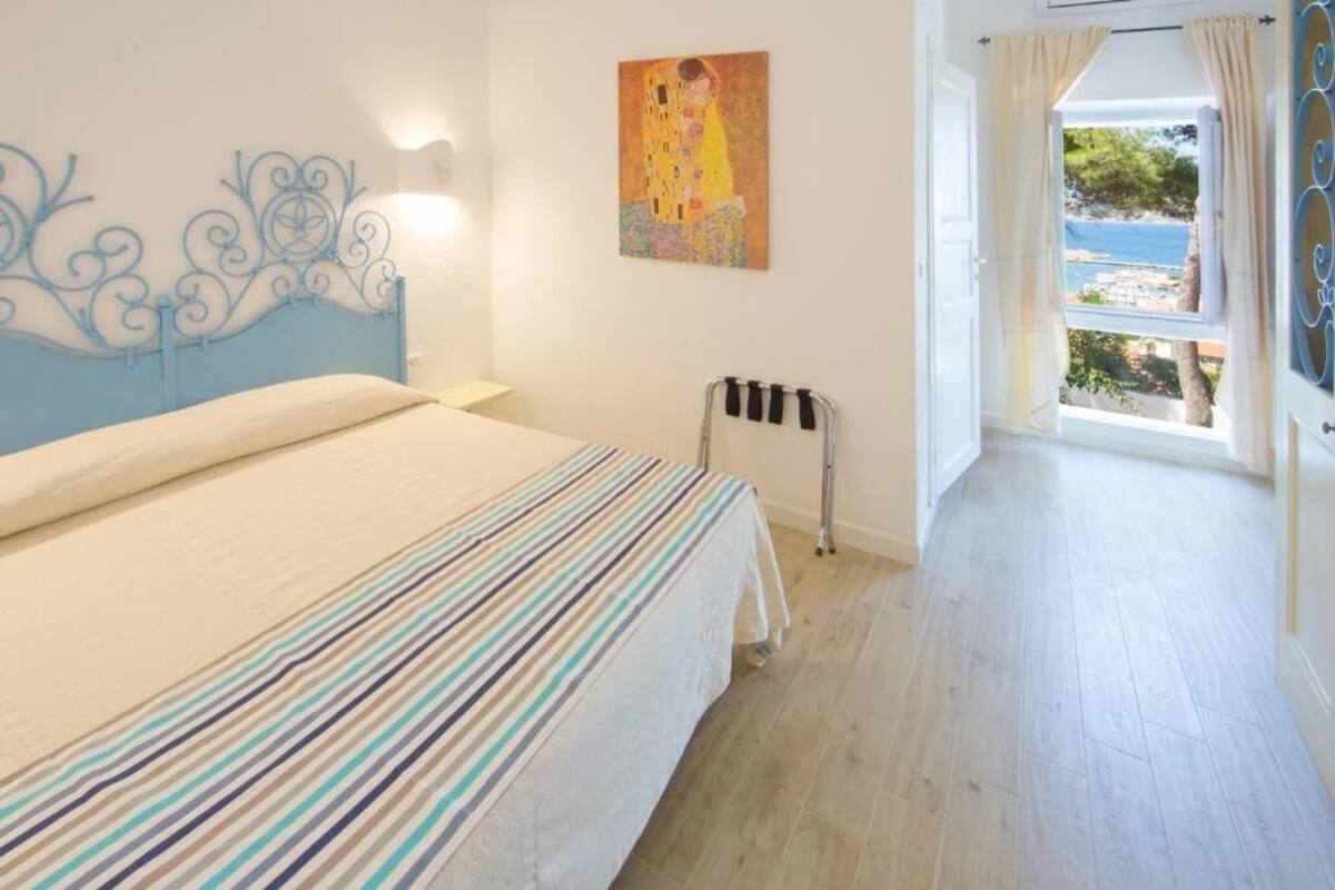 A bedroom with a bed, a radiator, a painting, and a view of the sea.