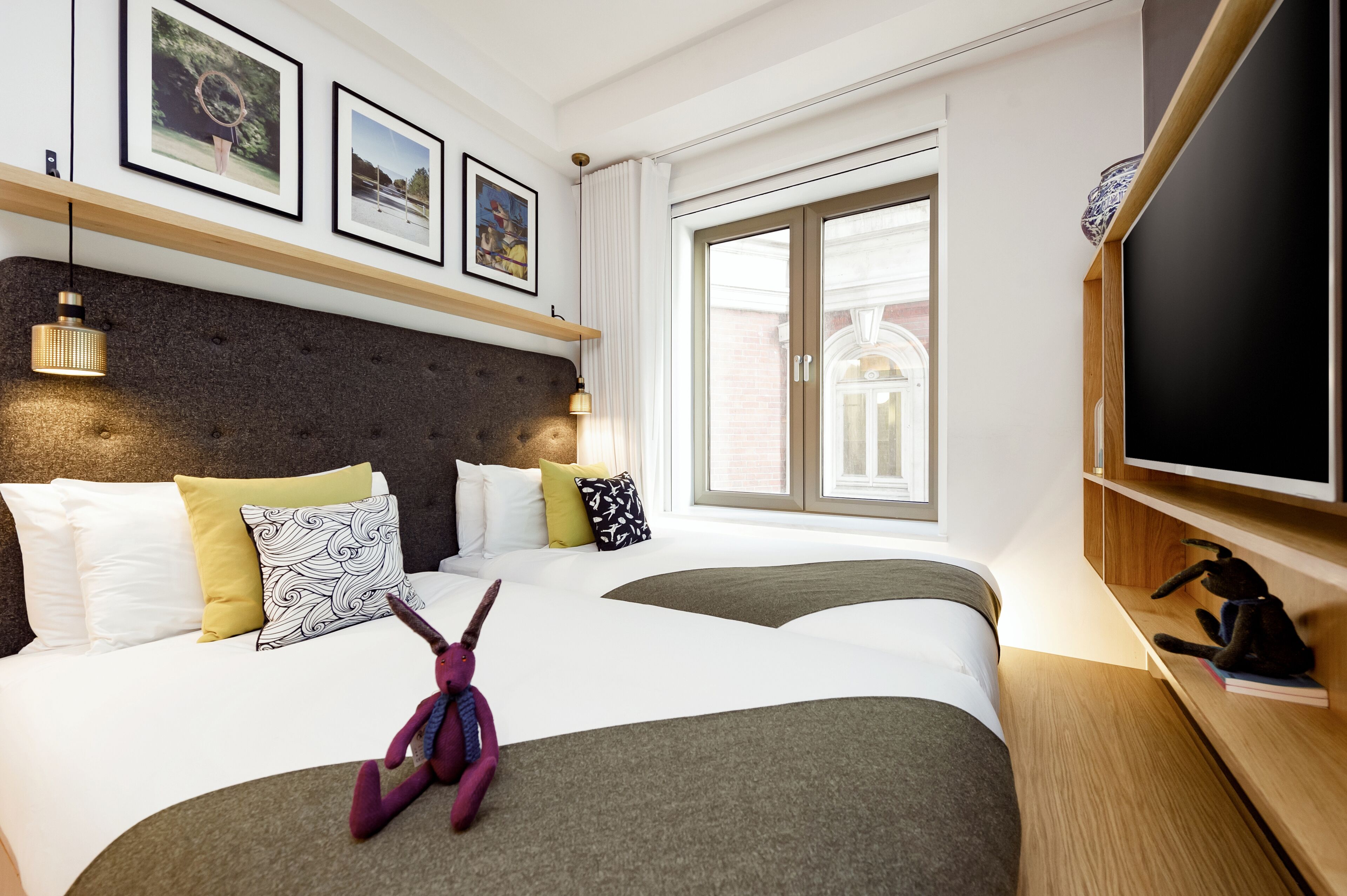 Ett hotellrum med två sängar, ett stort fönster, en platt-TV och inramade tavlor på väggen.