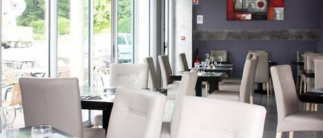 Un restaurante con sillas de cuero blanco, mesas dispuestas con copas de vino y platos, y amplios ventanales que ofrecen vistas al exterior.