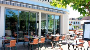 Een terras van een restaurant met tafels en stoelen, grote ramen en een gebouw met een bord waarop 