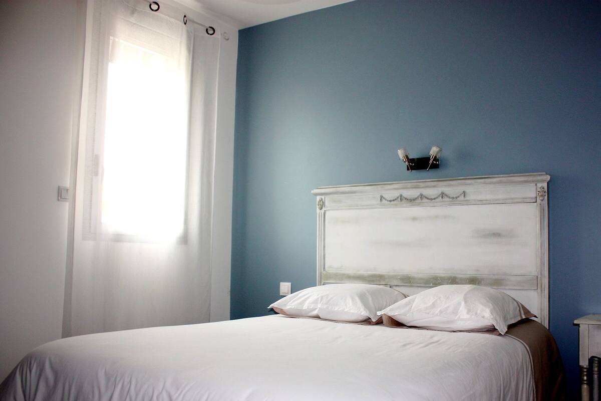 Een slaapkamer met een bed, wit beddengoed, een blauwe wand en een raam met gordijnen.