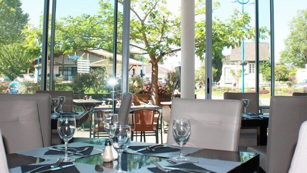 Een terras met eettafels voor twee personen, helder servies en uitzicht op de groene omgeving en gebouwen.