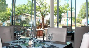 Un espace de restauration en plein air avec des tables pour deux personnes, de la vaisselle transparente et une vue sur la végétation et les bâtiments environnants.