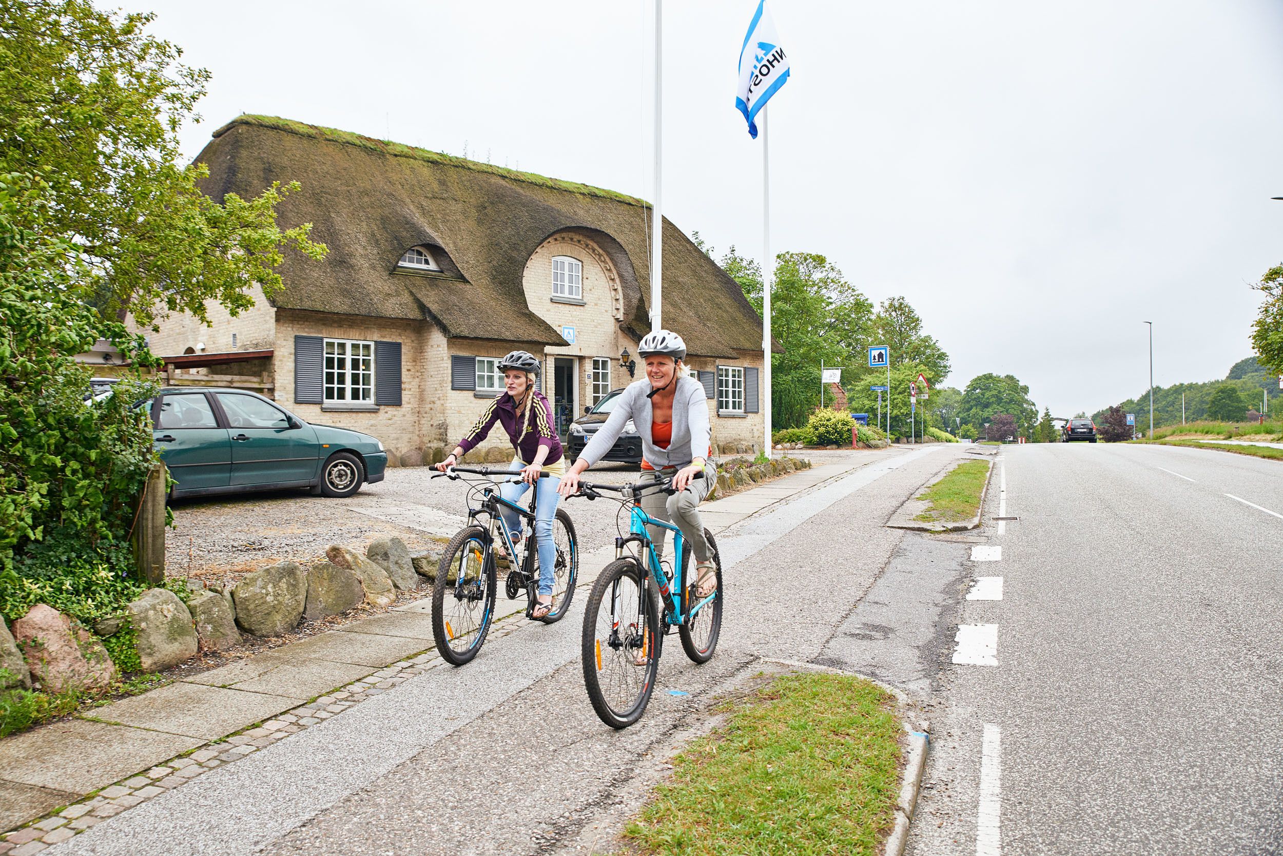 To cyklister på en belagt sti ved siden af en vej med en traditionel bygning med stråtækt tag.
