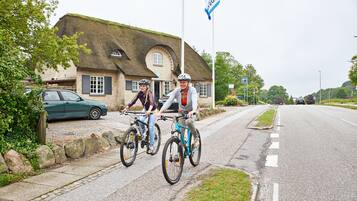To cyklister på en belagt sti ved siden af en vej med en traditionel bygning med stråtækt tag.