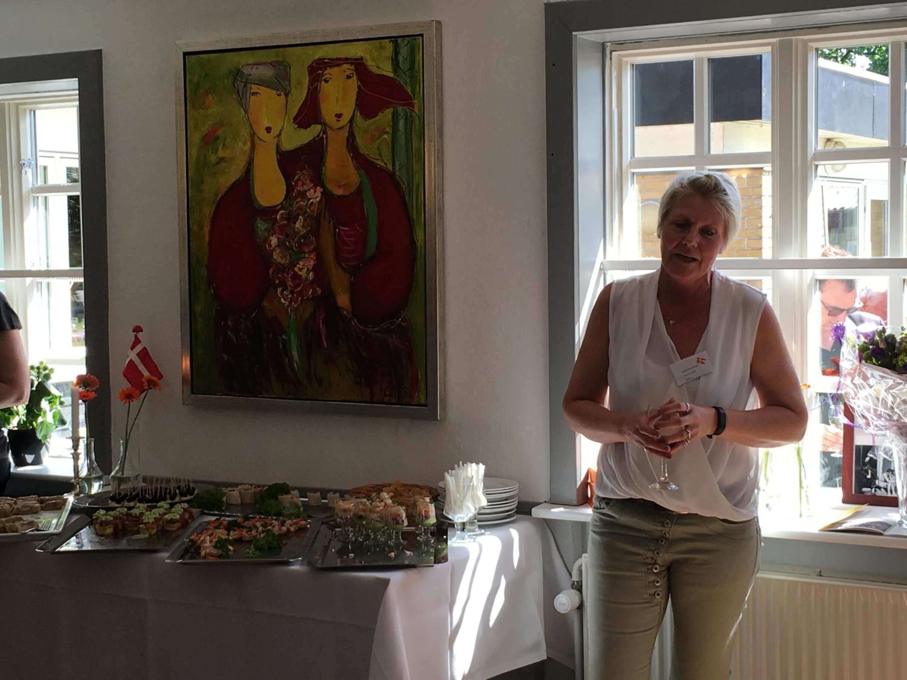 A room with a table set for a meal, a painting on the wall, and a window with a view of buildings.
