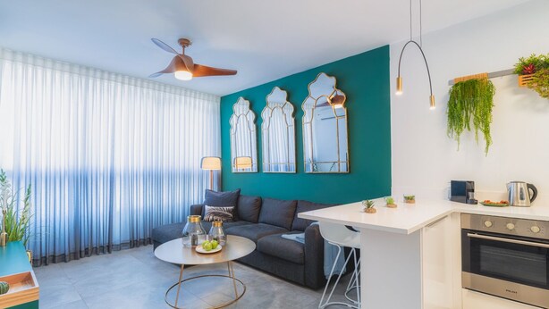 A modern living room with a sofa, a coffee table, and a kitchen area with a stove and hanging plants.