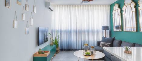 A modern living room with a green sofa, a coffee table, and a TV. There are curtains, a ceiling fan, and wall-mounted decorations.