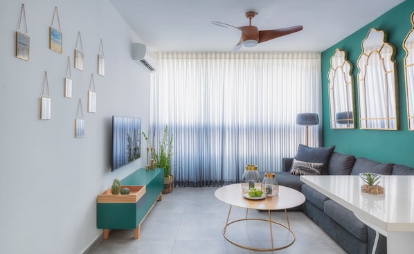 A modern living room with a green sofa, a coffee table, and a TV. There are curtains, a ceiling fan, and wall-mounted decorations.