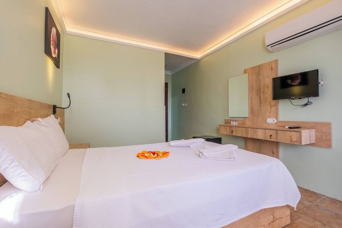 A hotel room with a bed, a wooden headboard, a flat-screen TV, a wooden vanity with a mirror, and a wall-mounted air conditioner.