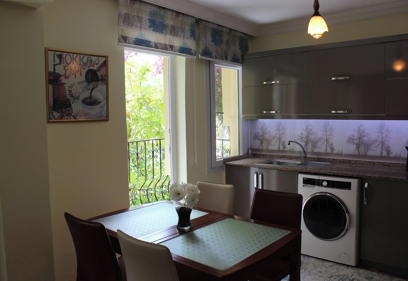 Cocina compacta con lavadora integrada, una mesa de comedor con sillas y ventana con vistas a zonas verdes.