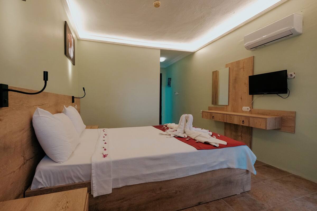 A neatly made bed with white linens and a red blanket, a wooden headboard, a bedside table, a wall-mounted TV, and an air conditioning unit.