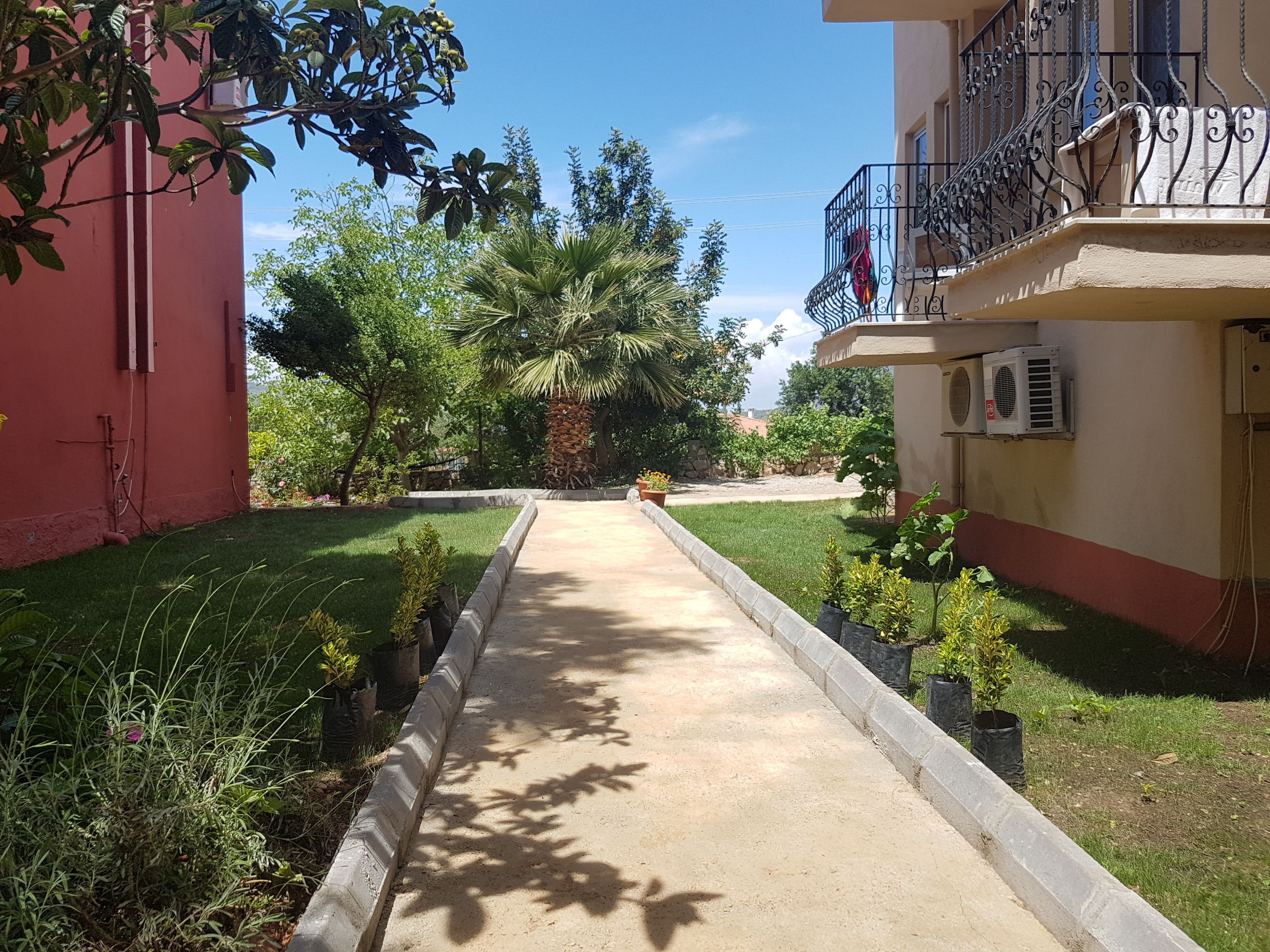A pathway leading to a building with a balcony, surrounded by greenery and a clear sky.