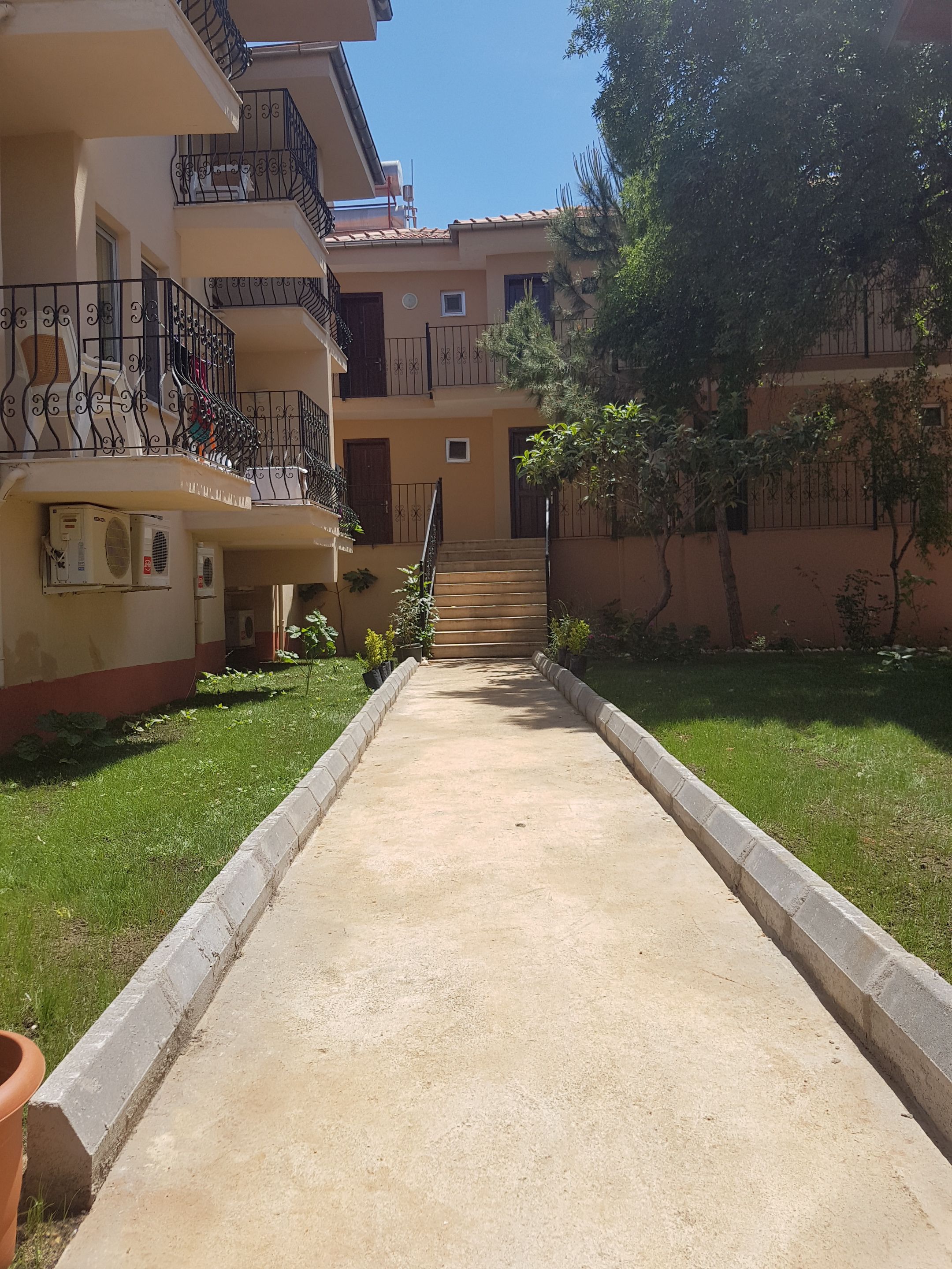 Un edificio residencial de varios pisos con balcones, un camino de hormigón y una pequeña zona ajardinada.