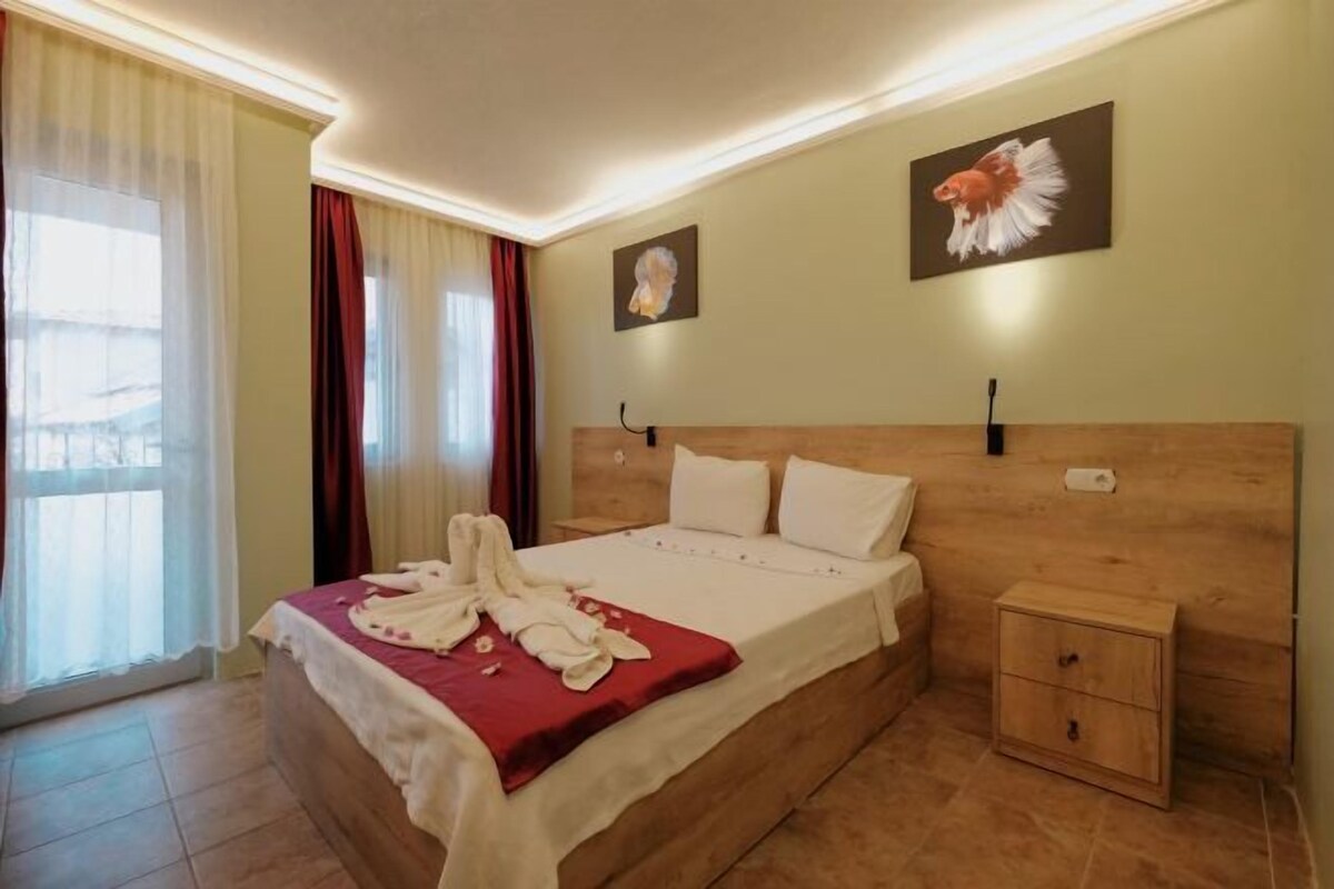 Habitación de hotel con cabecera de madera, cama con ropa de cama blanca, mesita de noche y un pequeño armario de madera.
