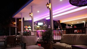 Un espacio al aire libre con sillas y mesas acolchadas, una zona de bar con televisor e iluminación ambiental.