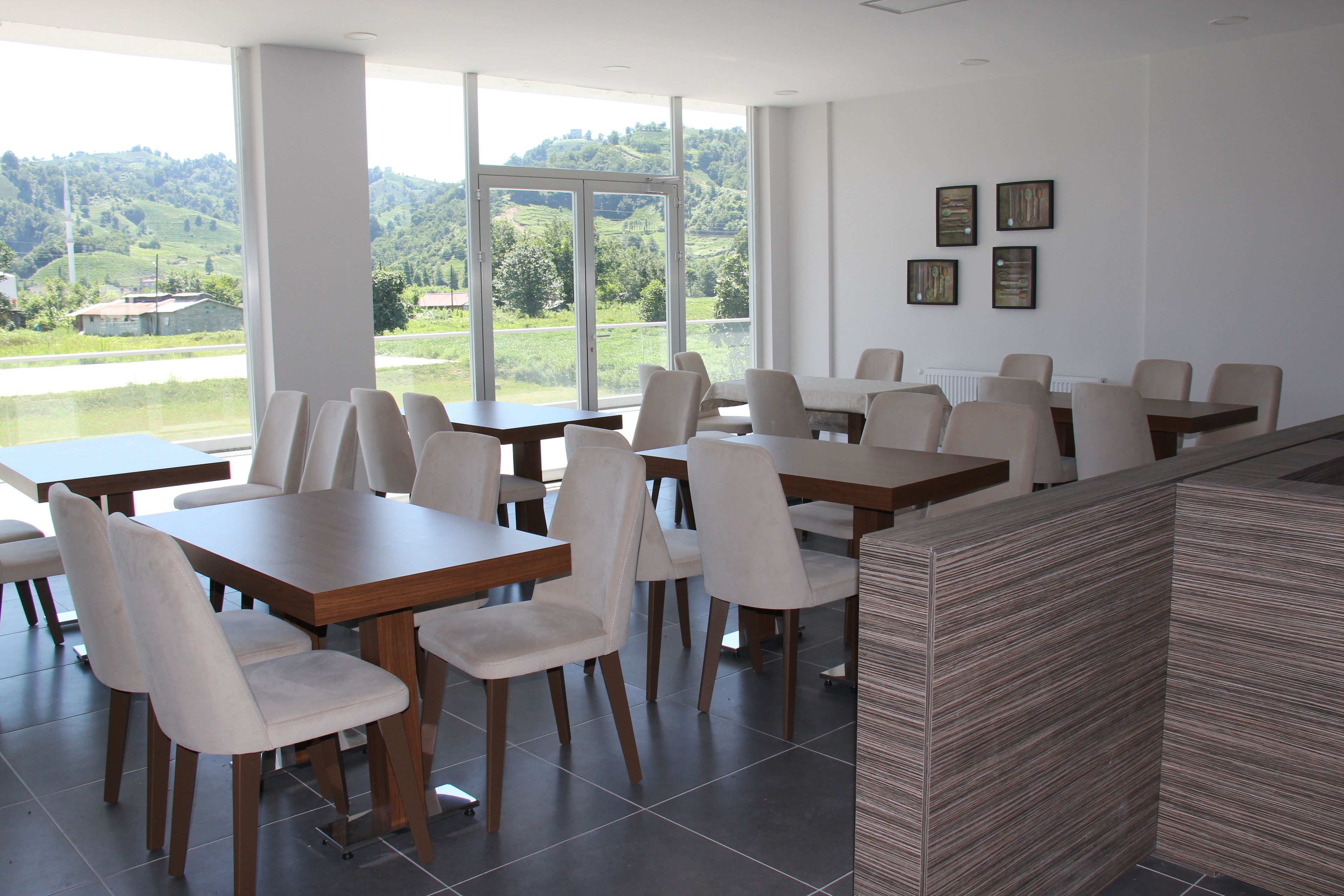 Un espace de restauration moderne, doté de plusieurs tables et chaises, de grandes fenêtres offrant une vue sur la verdure et les bâtiments, ainsi que d’œuvres d’art accrochées aux murs.