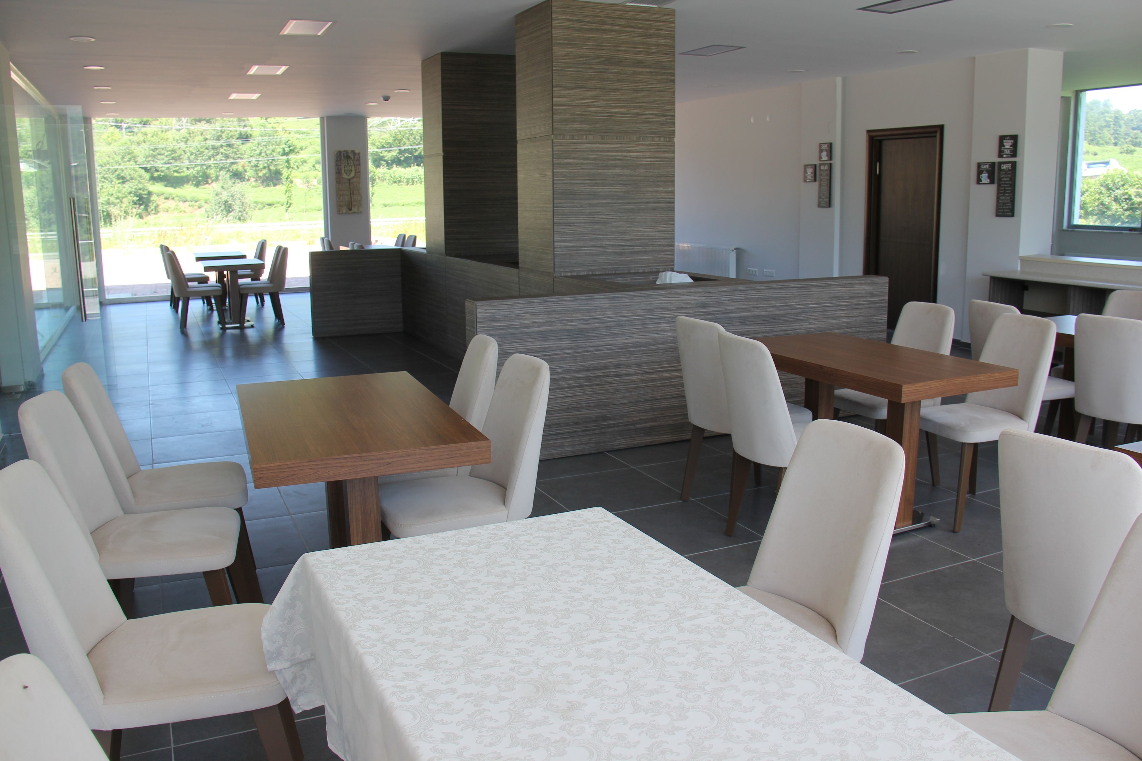 A modern restaurant with a central bar, multiple seating areas, and a view of greenery outside.