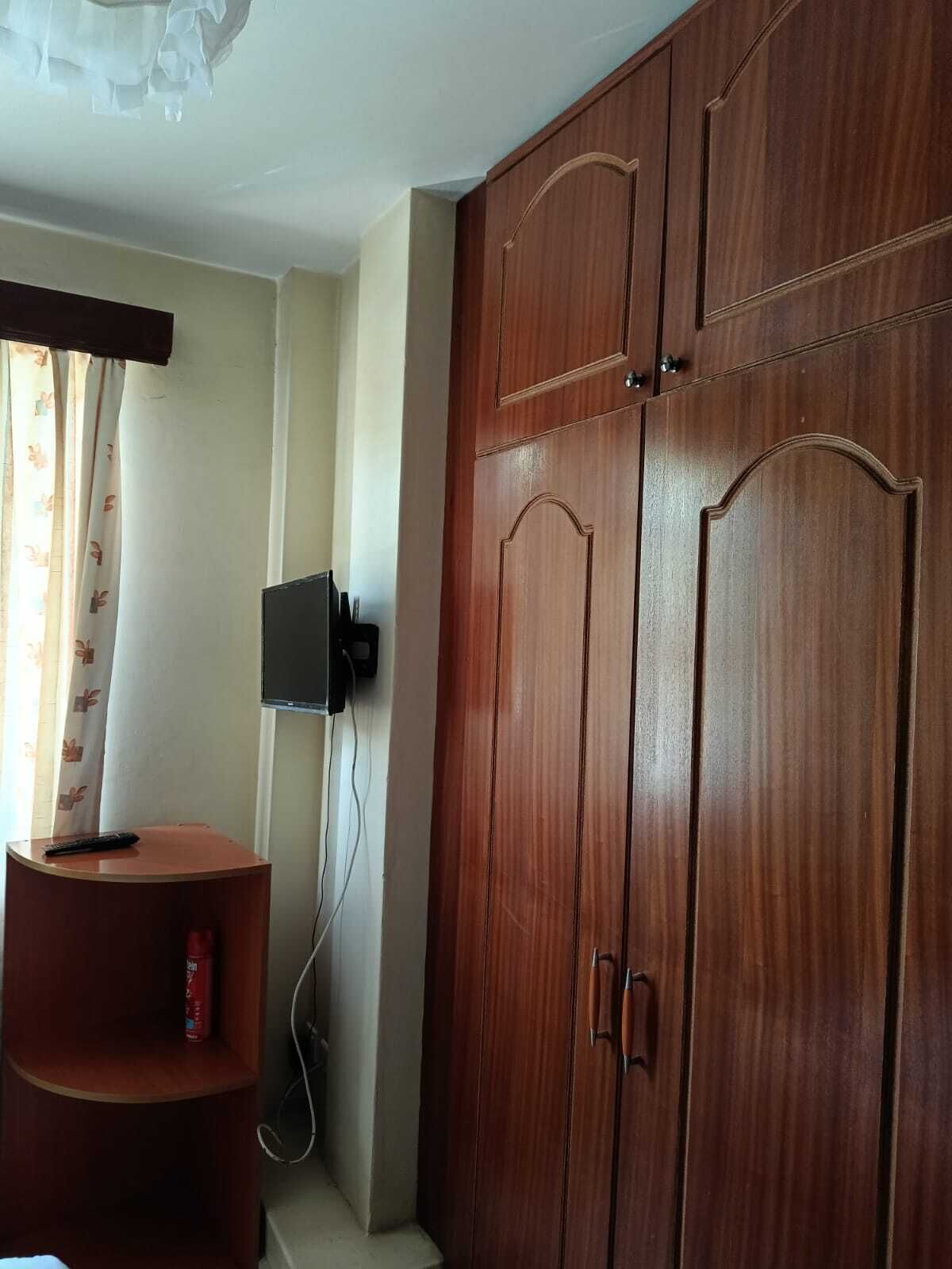 Une pièce avec une armoire en bois, un téléviseur fixé au mur et une petite table en bois sur laquelle se trouve une télécommande.