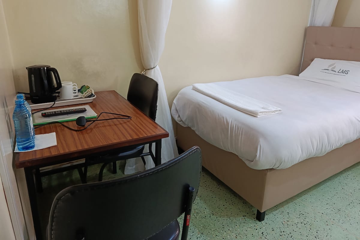 A hotel room with a bed, a desk with a kettle, a water bottle, and a chair.