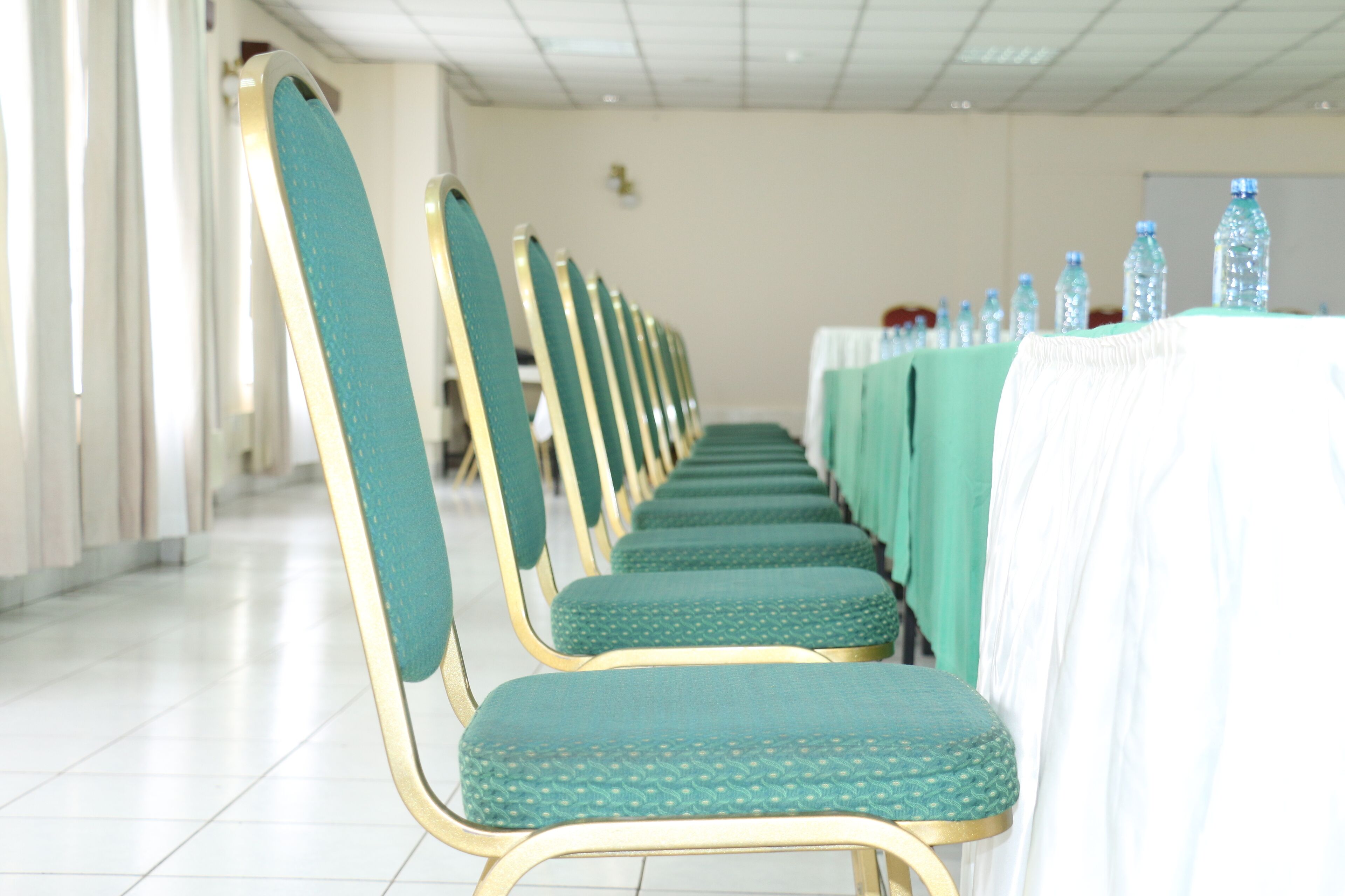 Rows of teal chairs with gold frames, a long table with a white tablecloth, and water bottles on the table.
