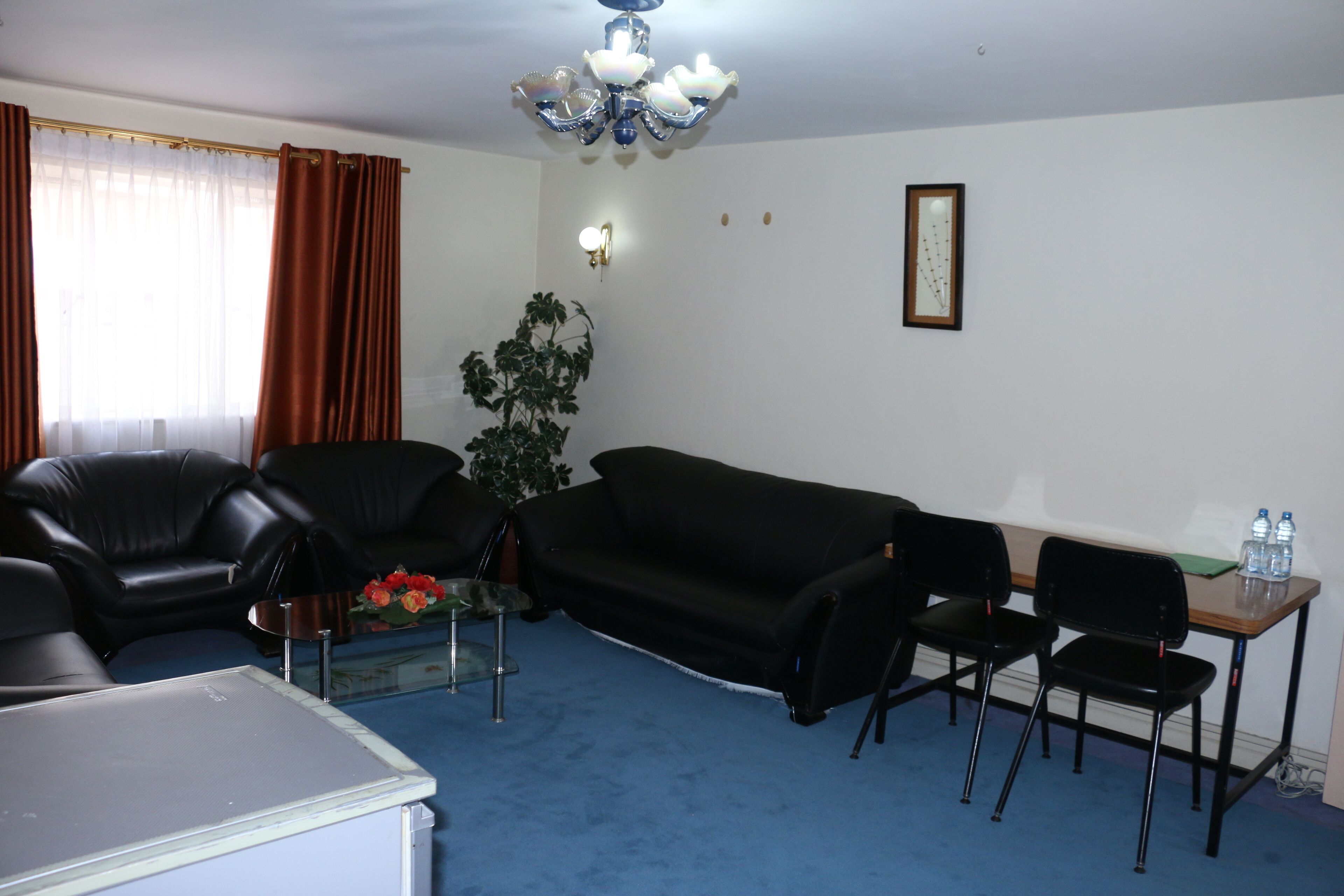 A living room with a black sofa, two black chairs, a glass coffee table with flowers, a small table with water bottles, a potted plant, and a chandelier.
