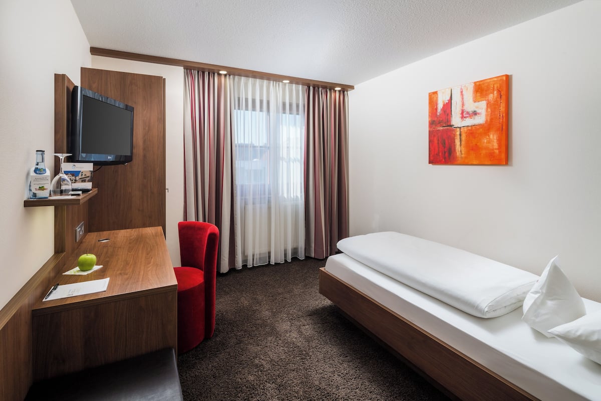 A hotel room with a wooden desk, a television, a bed, a red chair, and a painting on the wall.