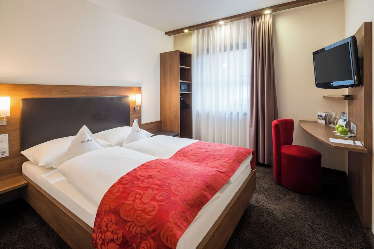 A hotel room with a large bed, a TV mounted on the wall, a desk with a chair, and a wardrobe.