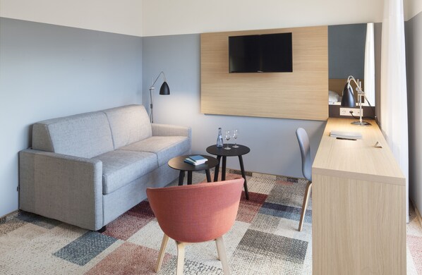 A modern hotel room with a grey sofa, a red armchair, a small round table with a book and glasses, a desk with a lamp, and a mounted TV.