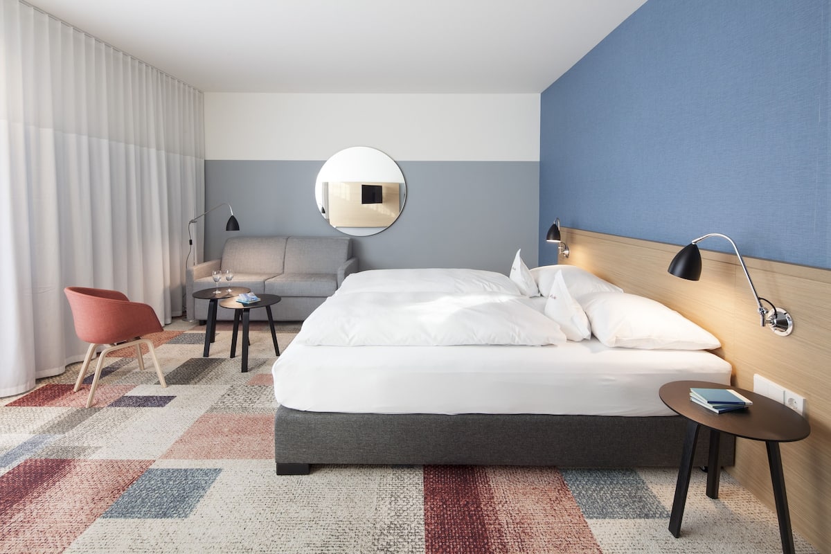A modern hotel room with a large bed, a round mirror, a red chair, a small table, and a blue wall.