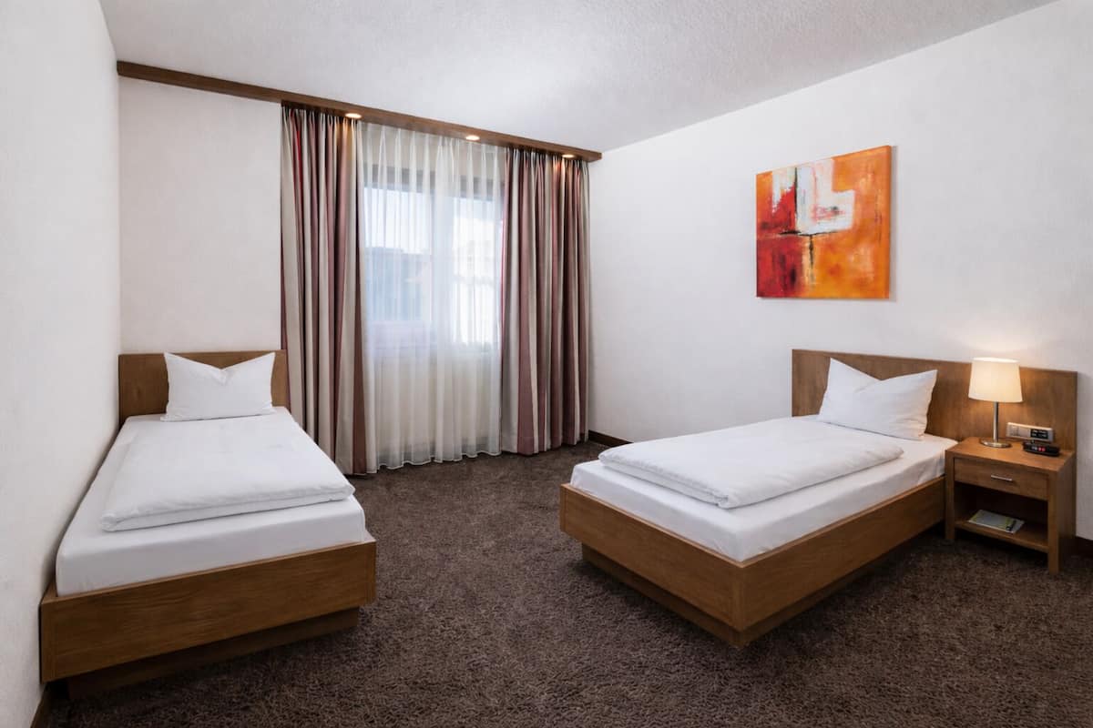 A hotel room with two single beds, a nightstand with a lamp, a painting on the wall, and a window with curtains.
