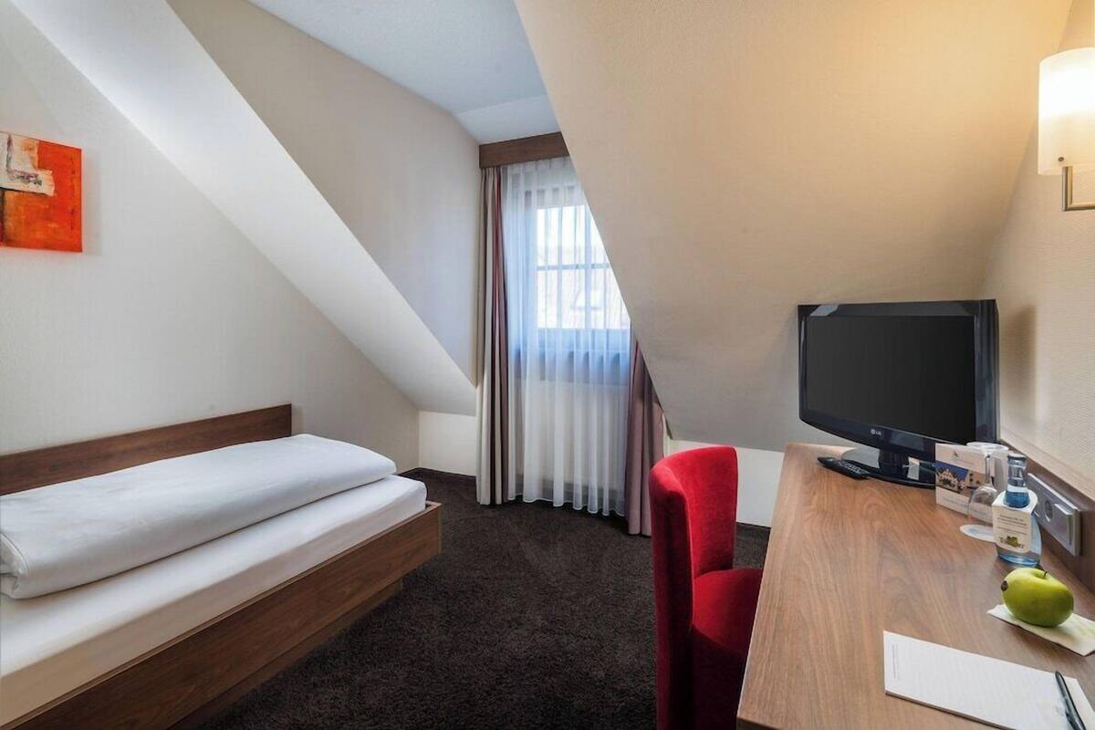 A hotel room with a bed, a desk with a TV, a chair, and a window with curtains.